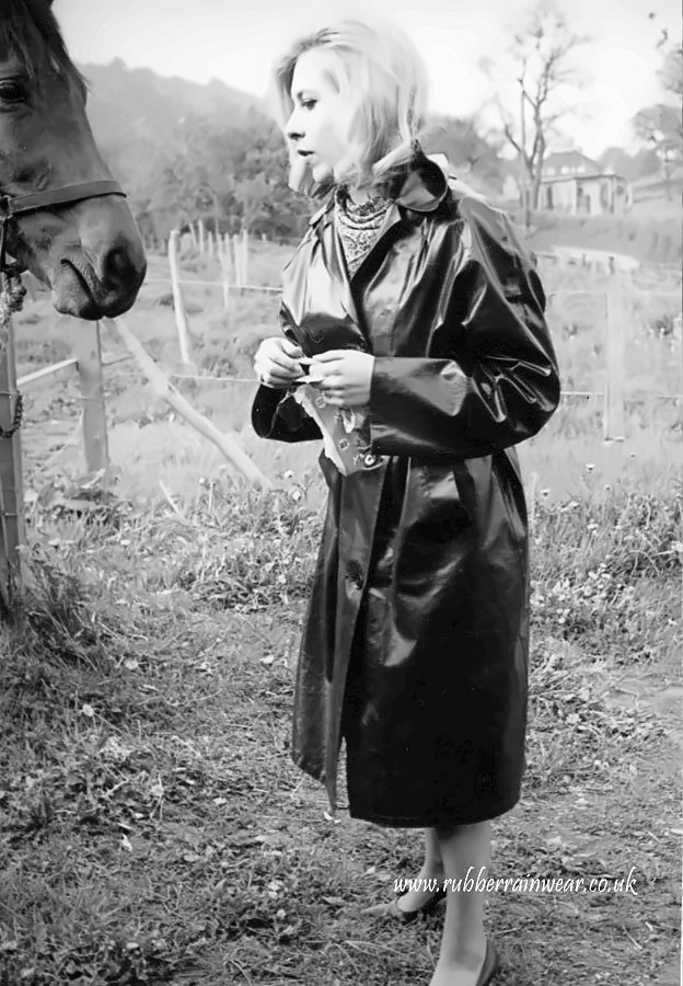 Woman with a blonde bob in a classic black rubberised mackintosh standing by a paddock fence, a horse looking on.