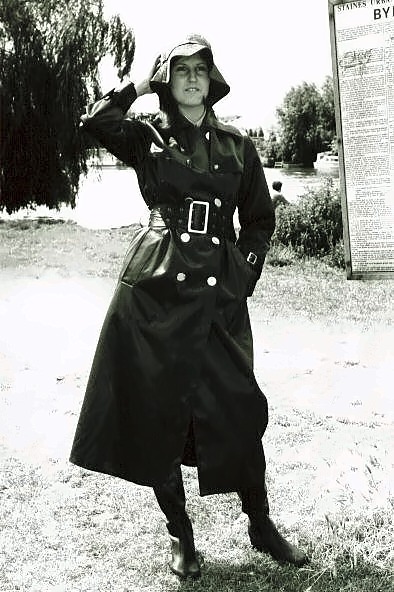 Woman in a hooded, double-breasted black belted mackintosh and boots saluting on a lawn.
