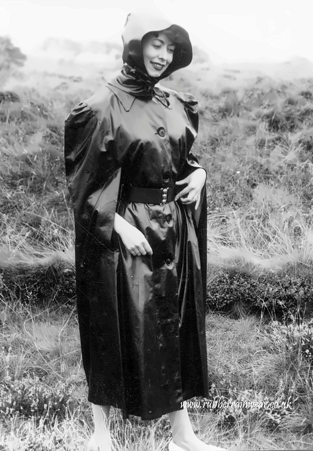 Woman in a full-length hooded black rubberised mackintosh with full puffed sleeves, buttoned and belted, standing on open heathland.