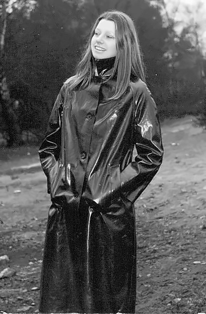 Smiling young woman in a long black rubber mackintosh with hands in pockets, on a woodland path.