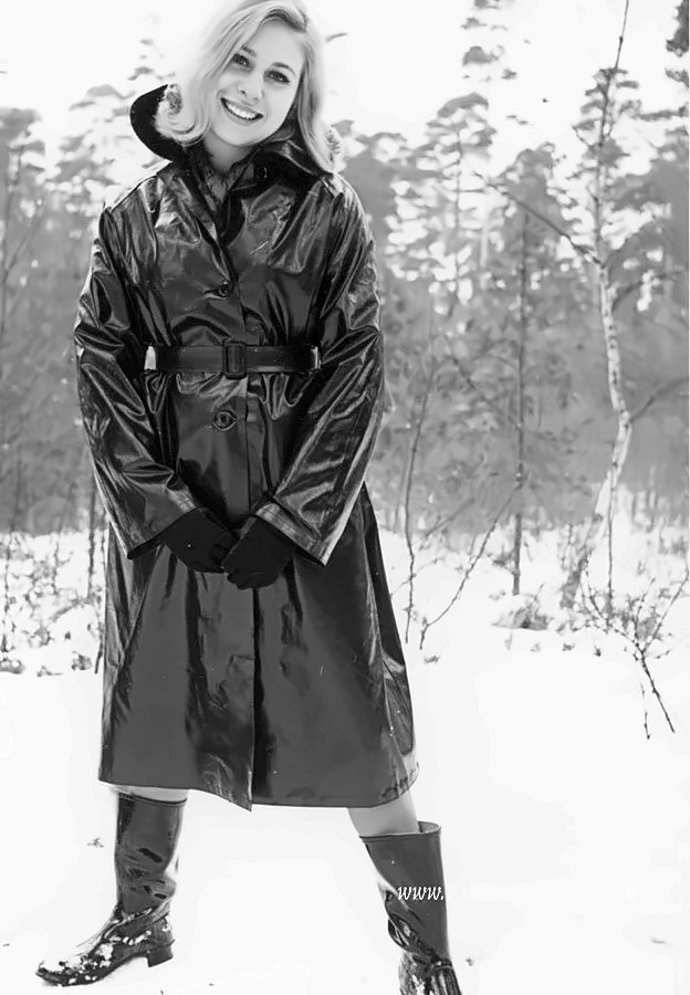 Smiling woman in a belted black mackintosh and rubber boots standing in snow among birch trees.