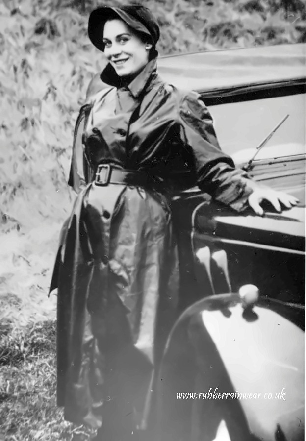 Woman in a cap and long belted black mackintosh leaning against a vintage motor car.
