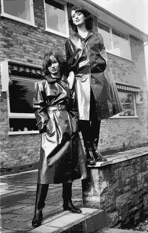 Two women in long belted mackintoshes and boots posing outside a brick house.